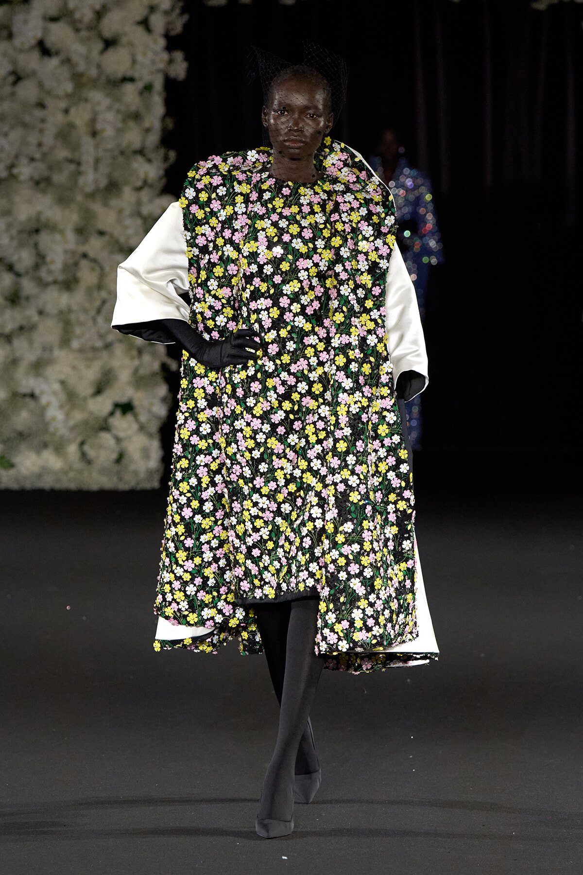 Model on a runway wearing a floor-length black floral dress with pink, yellow, and white flowers, white voluminous sleeves, black gloves, and a black net headpiece.