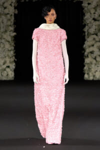 Model walking the runway in a floor-length pink sequin gown with a white oversized collar and long pale sleeves, against a dark backdrop with floral decor edges.]','Model on a fashion show runway wearing a textured pink sequined dress with a white collar and long white sleeves.