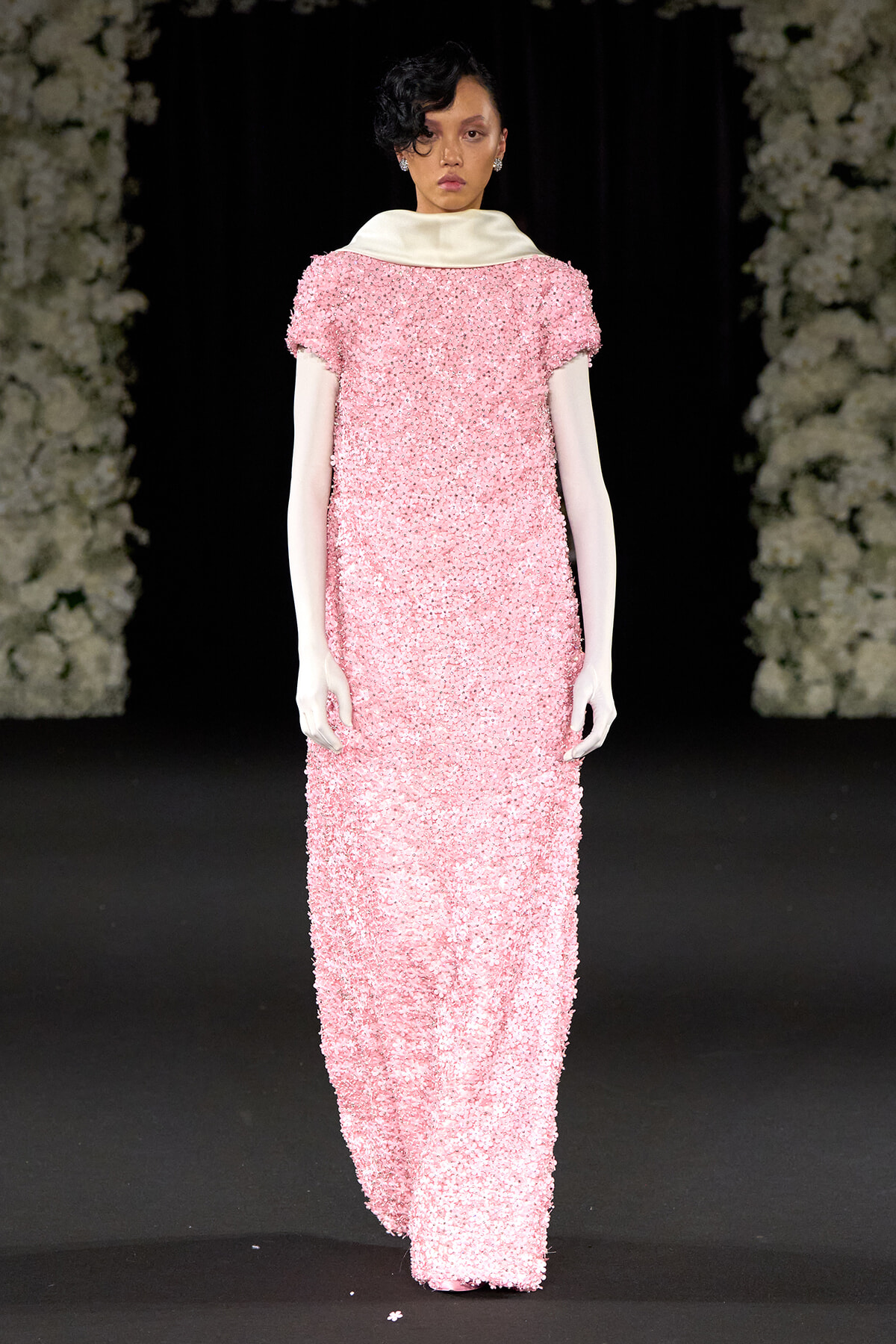 Model walking the runway in a floor-length pink sequin gown with a white oversized collar and long pale sleeves, against a dark backdrop with floral decor edges.]','Model on a fashion show runway wearing a textured pink sequined dress with a white collar and long white sleeves.
