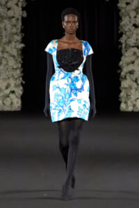 Runway model wearing a white blue floral mini dress with a black sequin bustier and bow, paired with long black gloves and tights on a dark catwalk