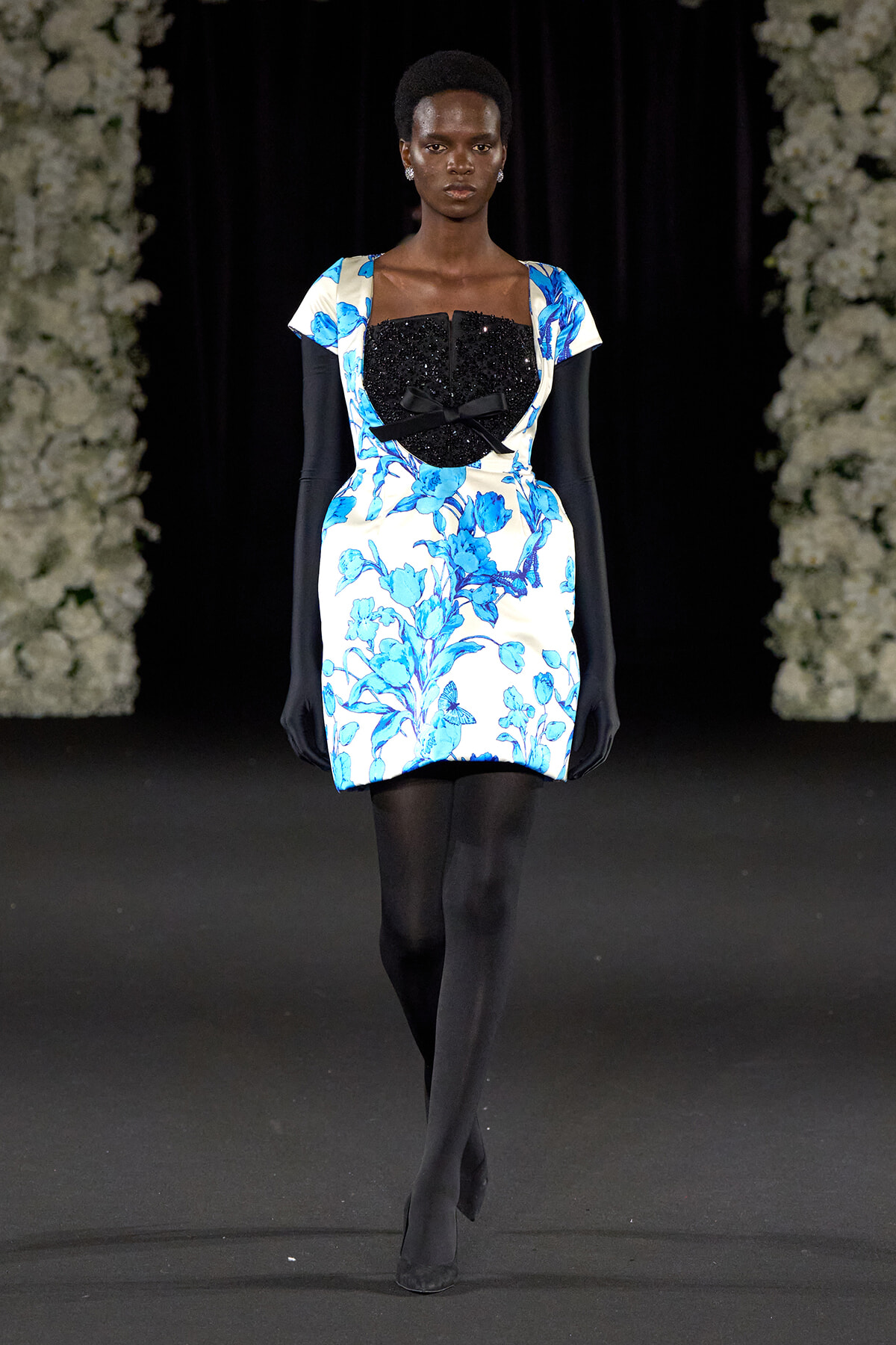 Runway model wearing a white blue floral mini dress with a black sequin bustier and bow, paired with long black gloves and tights on a dark catwalk