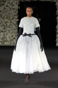 Model on a runway wearing a white beaded ball gown with a black ribbon belt, long black gloves, and a black net veil.