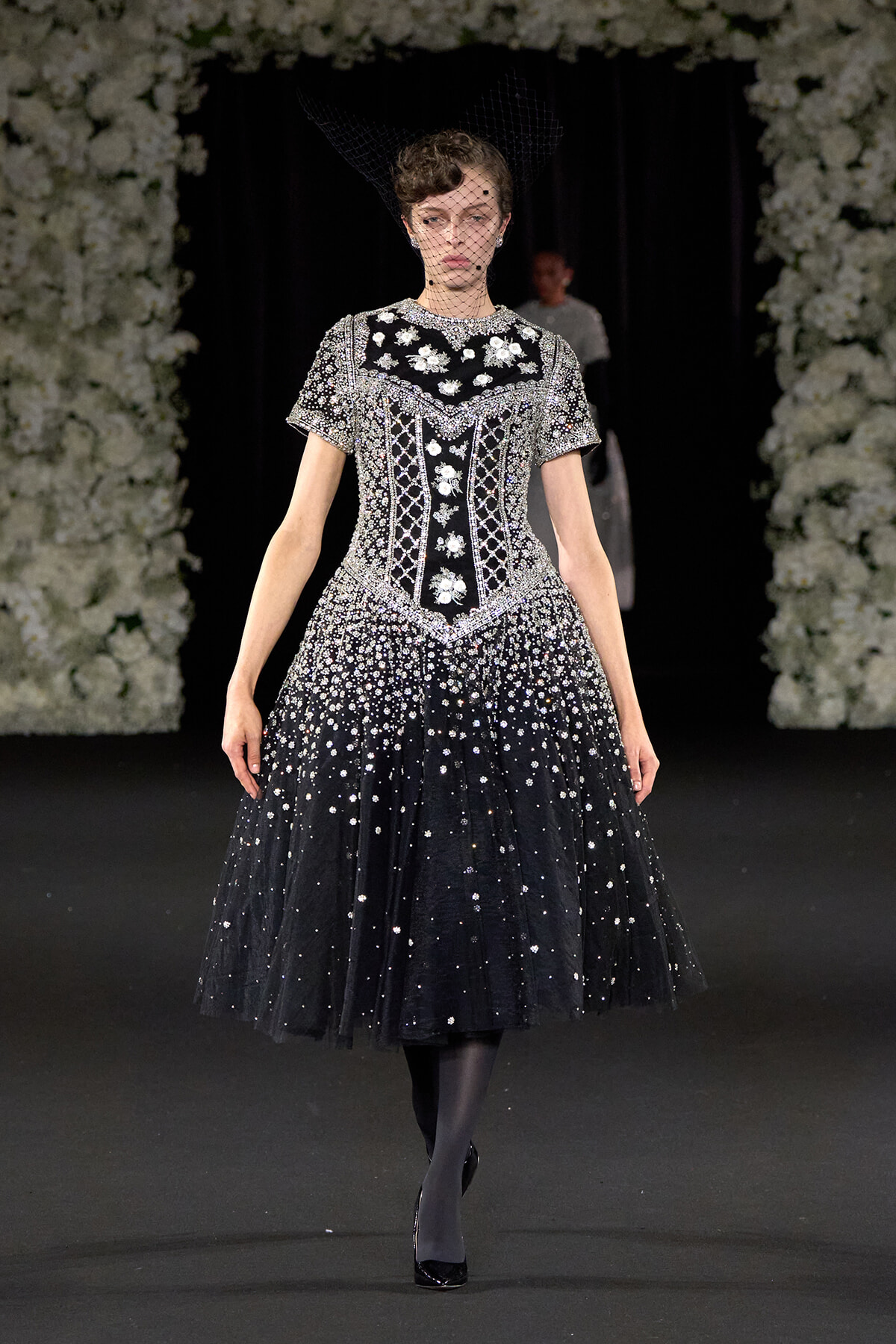 Model on a runway wearing a black, heavily embellished gown with silver beadwork and a net veil headpiece, walking the catwalk.