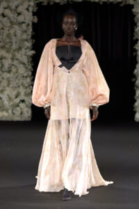 Runway model wearing a pale peach gown with voluminous sleeves, a black bodice, and sparkling details on a dark catwalk.