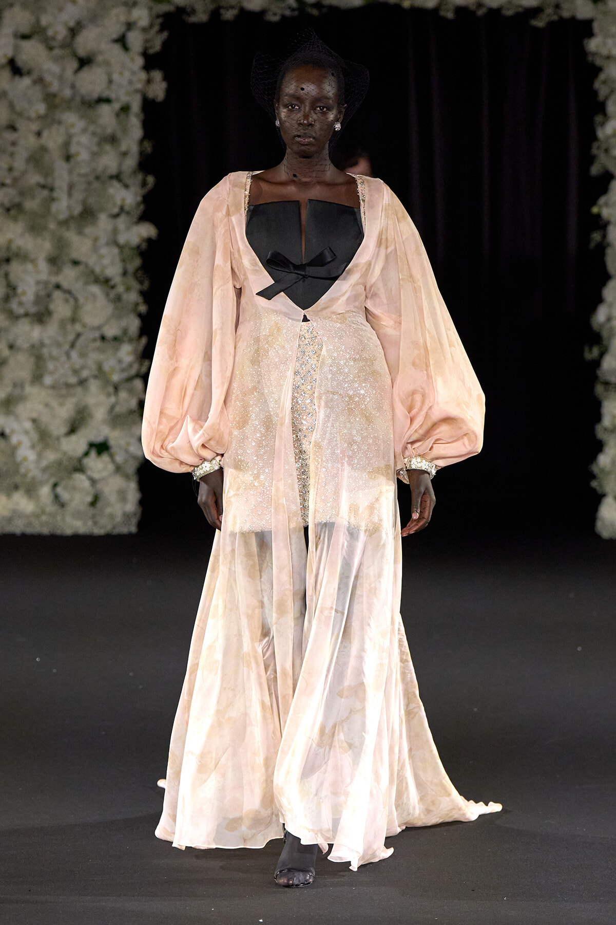 Runway model wearing a pale peach gown with voluminous sleeves, a black bodice, and sparkling details on a dark catwalk.