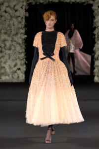 Model walking runway in a peach beaded ball gown with a black sequined bodice, black gloves, and a birdcage veil, accessorized with a black bow at the waist.