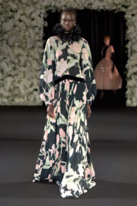 Model walking runway in a long floral gown with dark background and a black ruffled high collar, floral print in pink and green.