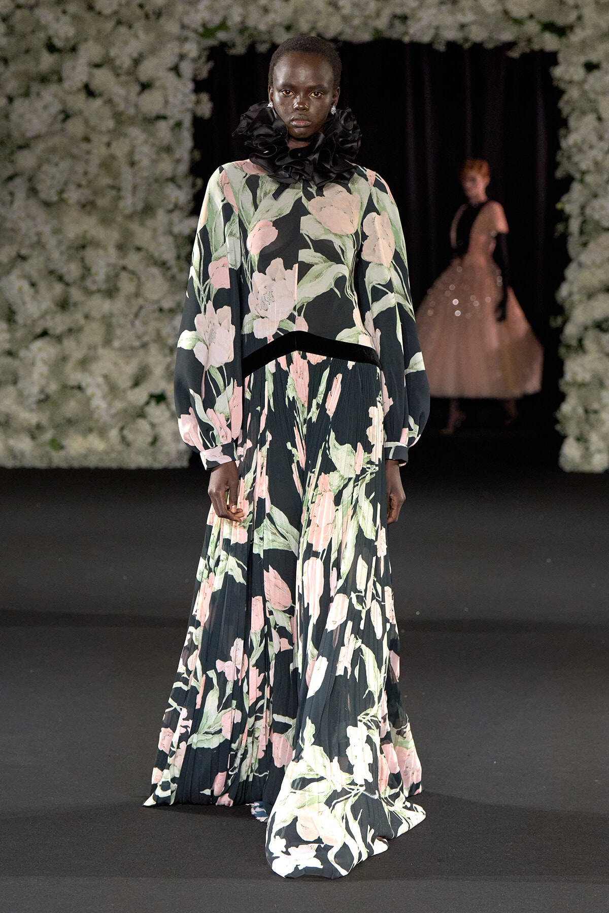 Model walking runway in a long floral gown with dark background and a black ruffled high collar, floral print in pink and green.