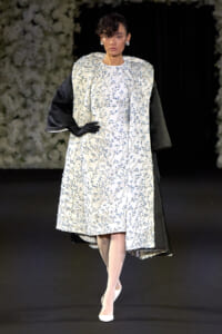 Model walks a runway wearing a white knee-length dress with blue floral embroidery and a contrasting pale cape over black-sleeved inner garment, white heels and gloves.