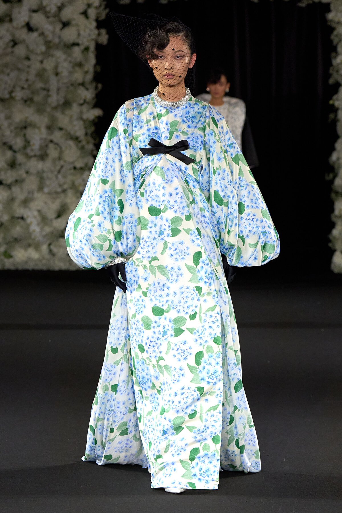 Model walking the runway in a long floral kimono-style gown with blue hydrangea pattern, green leaves, and a black bow at the waist, wearing a black mesh veil.