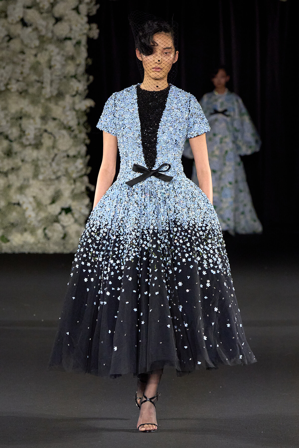 Model walking a runway in a two-tone evening gown: pale blue beaded bodice fading to black, with a black ribbon bow at the waist and a full tulle skirt adorned with white flowers.