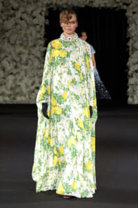 Model on a runway wearing a floor-length ivory gown with a yellow rose floral print, green leaves, and a jeweled high collar with a net veil and black gloves.