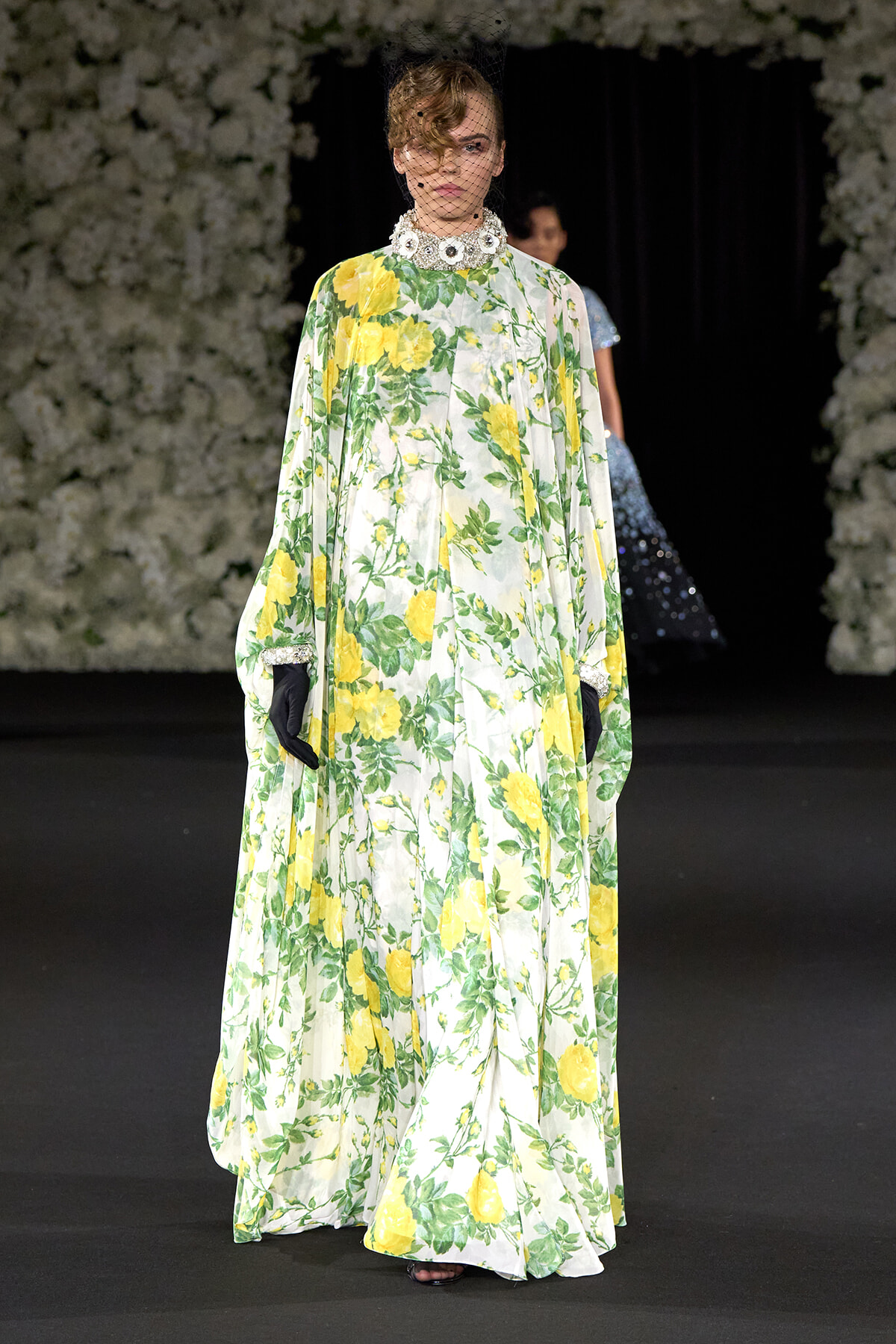 Model on a runway wearing a floor-length ivory gown with a yellow rose floral print, green leaves, and a jeweled high collar with a net veil and black gloves.