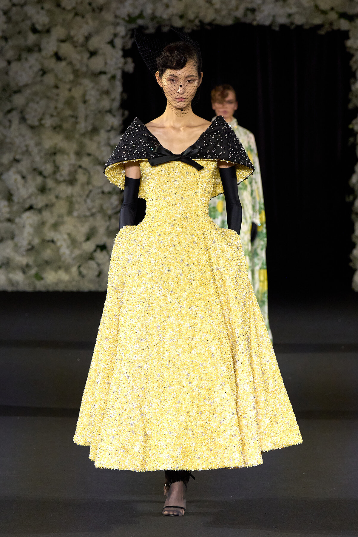 Model walking a runway in a sparkling yellow ball gown with a black sequined cape, black bow at the neckline, long black gloves, and a delicate veil.