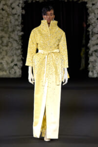 Model on a runway wearing a yellow, sequined coat with a tall collar and a satin belt, paired with pale yellow pants and white gloves.