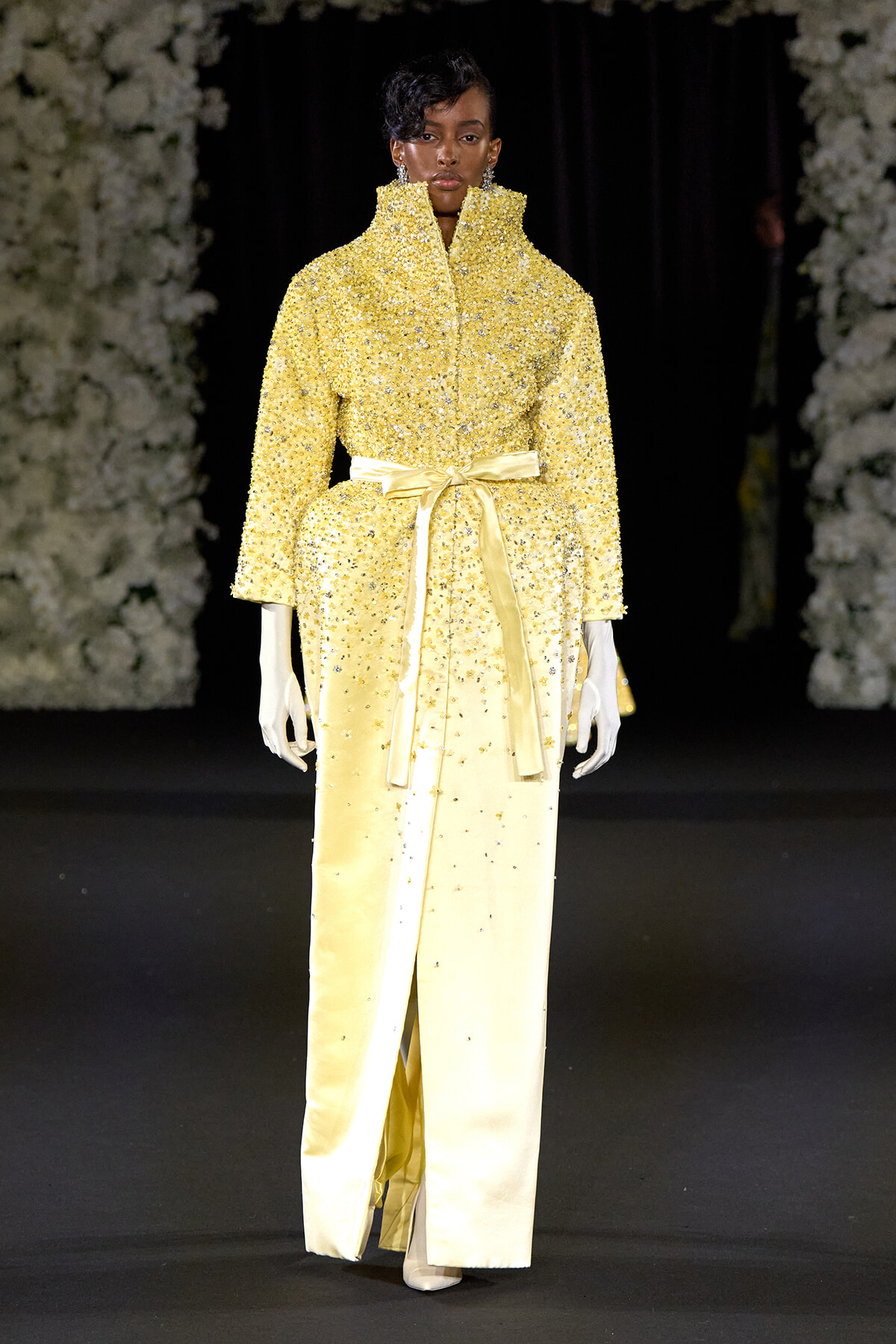 Model on a runway wearing a yellow, sequined coat with a tall collar and a satin belt, paired with pale yellow pants and white gloves.