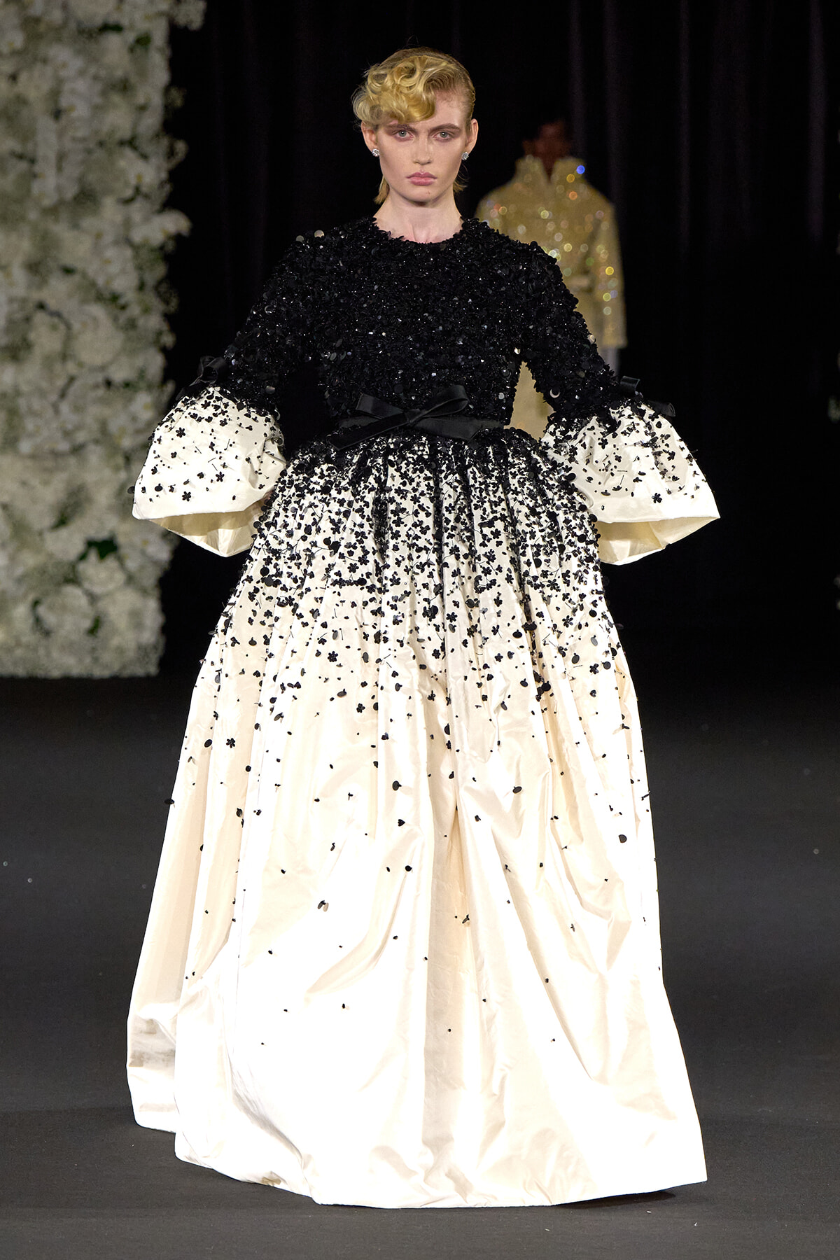 Runway model wearing a black sequined bodice with a full ivory skirt speckled with black beads and dramatic white puff sleeves, accented by a black sash at the waist.