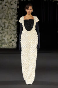 Model on a runway wearing a floor-length cream beaded gown with a black sequined bodice and bow, paired with black gloves and pearl earrings; floral backdrop to the left.
