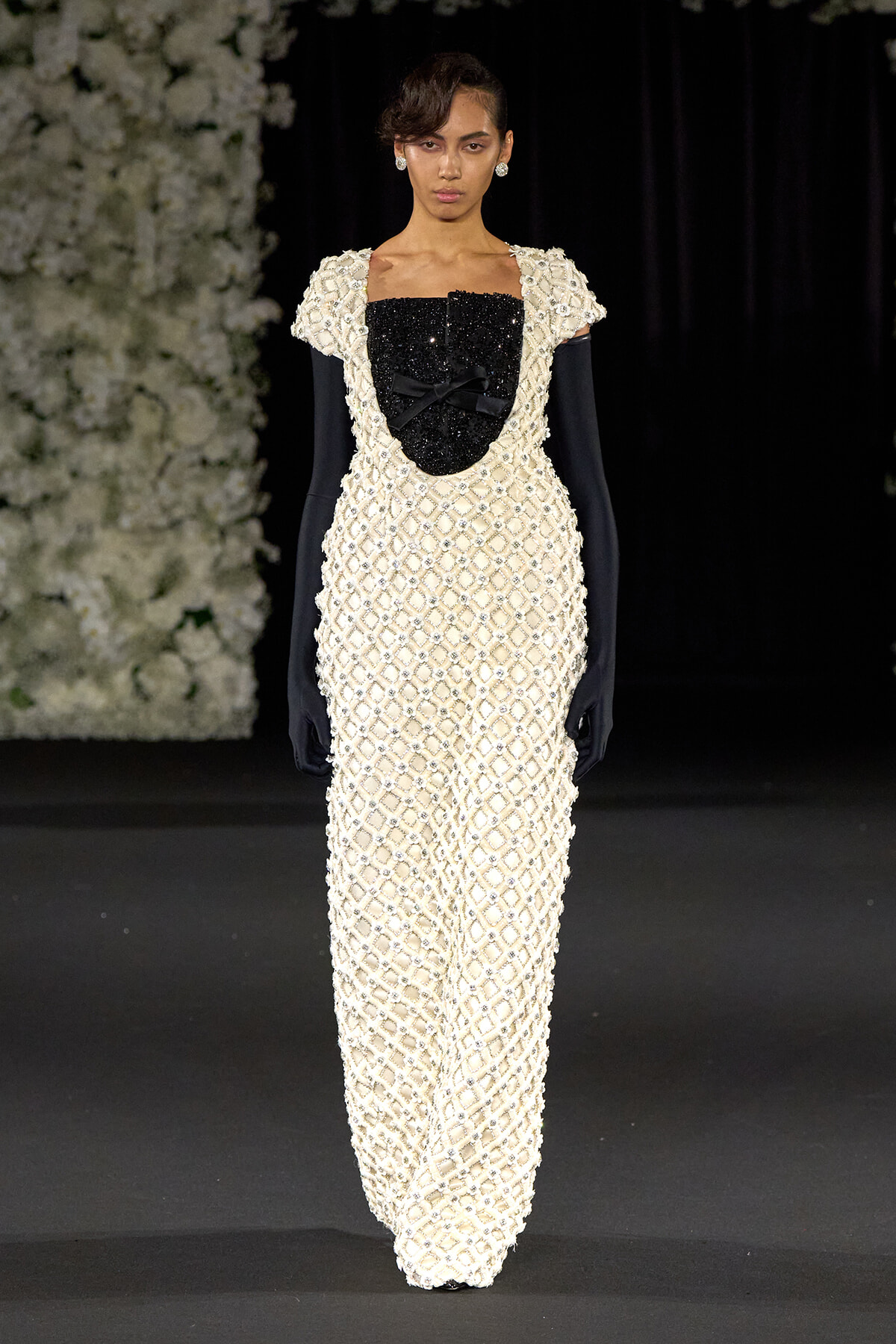 Model on a runway wearing a floor-length cream beaded gown with a black sequined bodice and bow, paired with black gloves and pearl earrings; floral backdrop to the left.