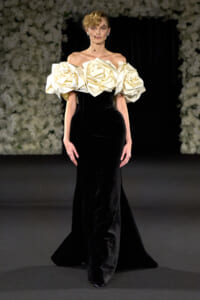 Model on runway wearing a black velvet gown with oversized cream rosette sleeves and a black net veil. Elegant haute couture look.