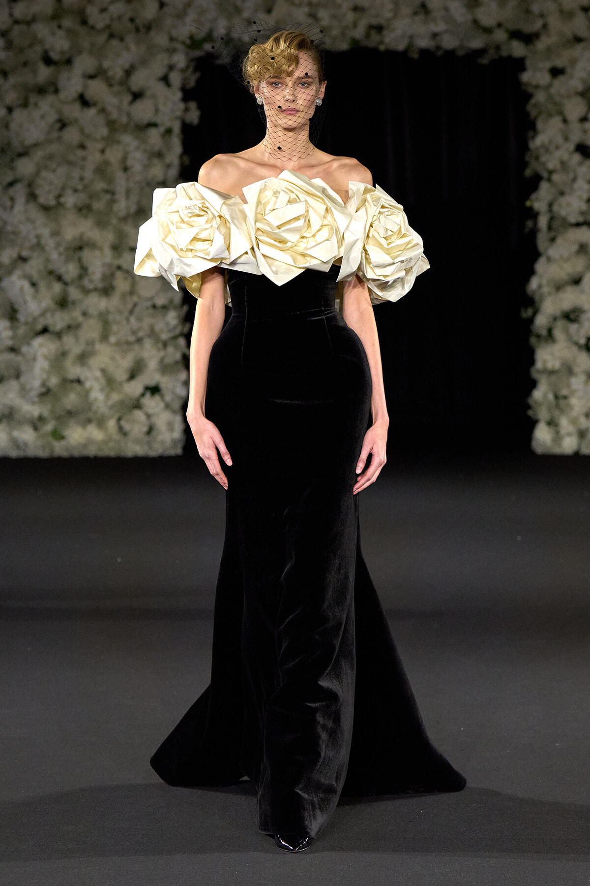 Model on runway wearing a black velvet gown with oversized cream rosette sleeves and a black net veil. Elegant haute couture look.