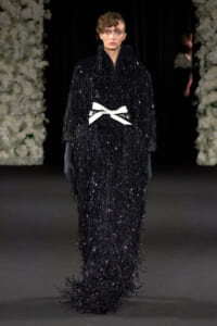 Model walks the runway in a floor-length black sequin fringe gown with a white bow belt and long gloves.