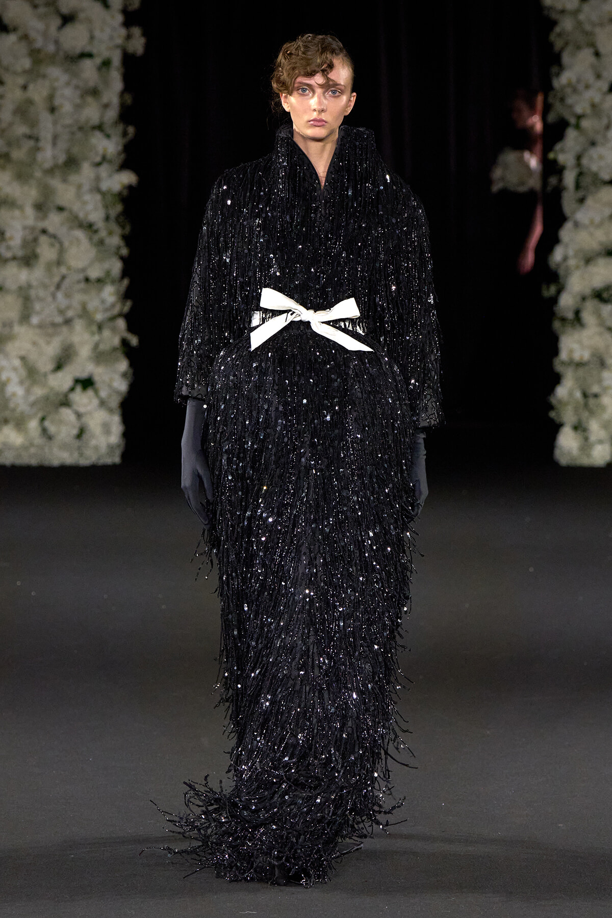Model walks the runway in a floor-length black sequin fringe gown with a white bow belt and long gloves.