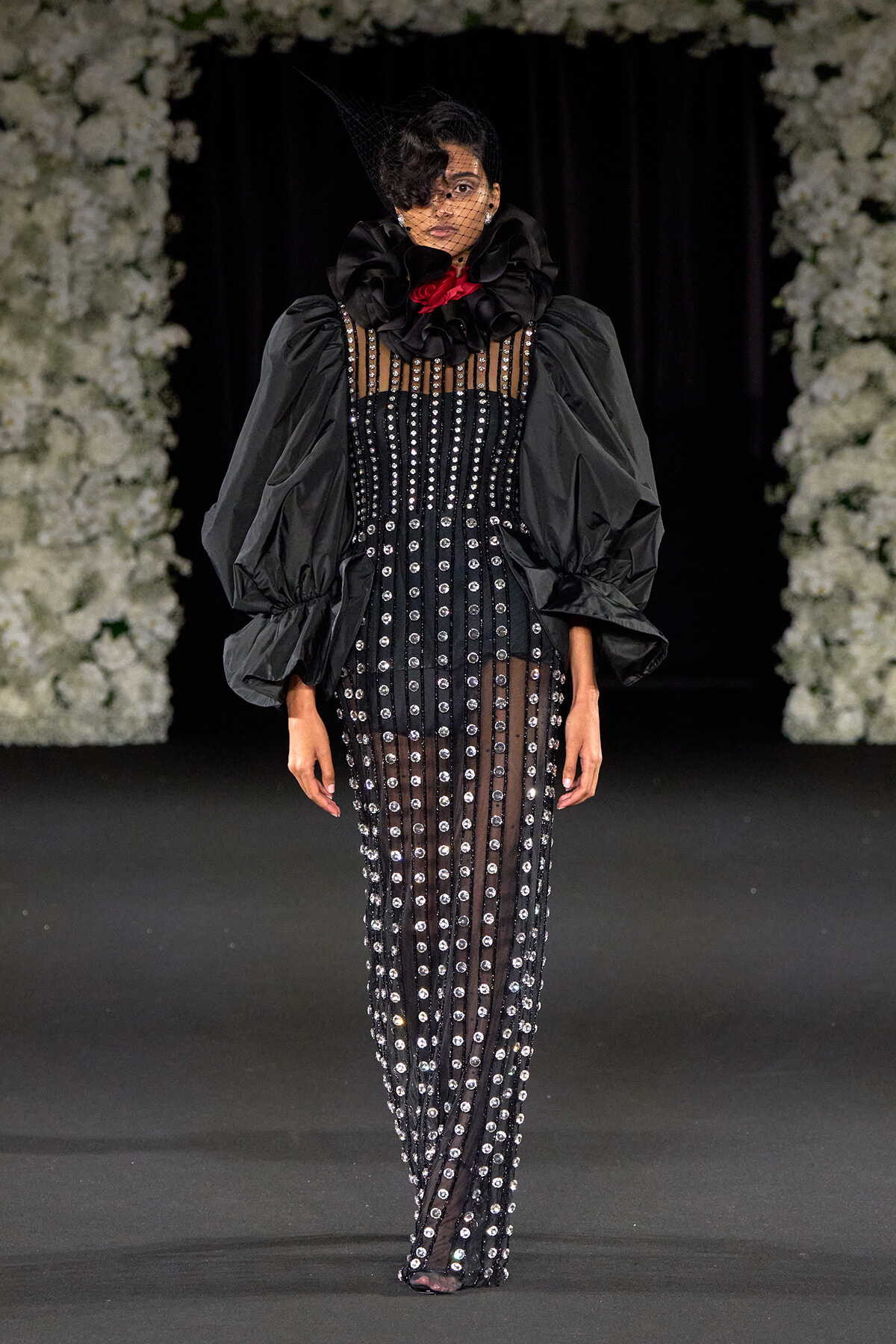 Runway model in a black sequined gown with sheer panels, oversized puff sleeves, and a ruffled neck collar with a red accent veil.