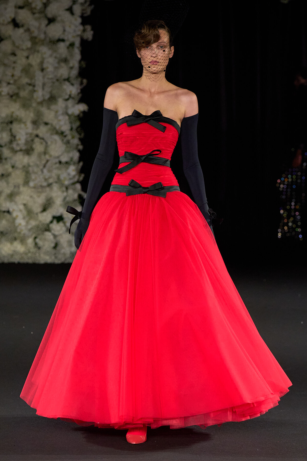 Model walks the runway in a vivid red off-shoulder ball gown with black bow accents, long black gloves, and a black net veil.
