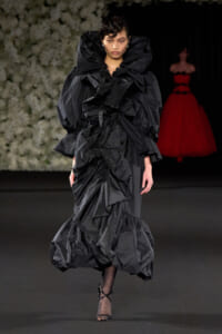 Model walking the runway in a dramatic black, ruffled gown with oversized collar and voluminous layers, dark backdrop behind.
