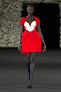 Model walking a fashion runway in a bright red, short dress with a white heart-shaped bodice and bow, paired with sheer black tights and black heels under a dark backdrop with floral arches.