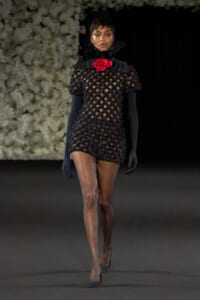 Model on a runway wearing a black lattice mini dress with sheer sleeves, a red rose at the neck, black gloves and a veil.