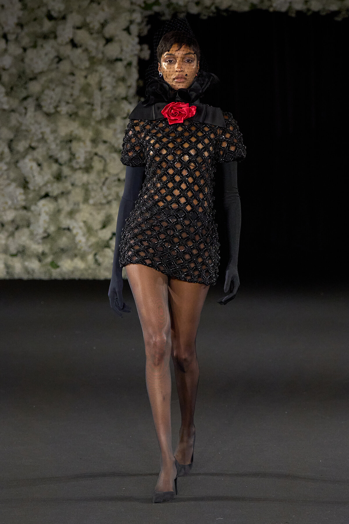 Model on a runway wearing a black lattice mini dress with sheer sleeves, a red rose at the neck, black gloves and a veil.