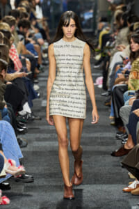 Model walking the runway in a sleeveless beige dress printed with black text, paired with brown ankle boots.
