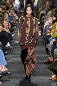Model walks a fashion runway in a brown patterned knit sweater with cutout sleeves and loose satin brown pants, audience on both sides watching.