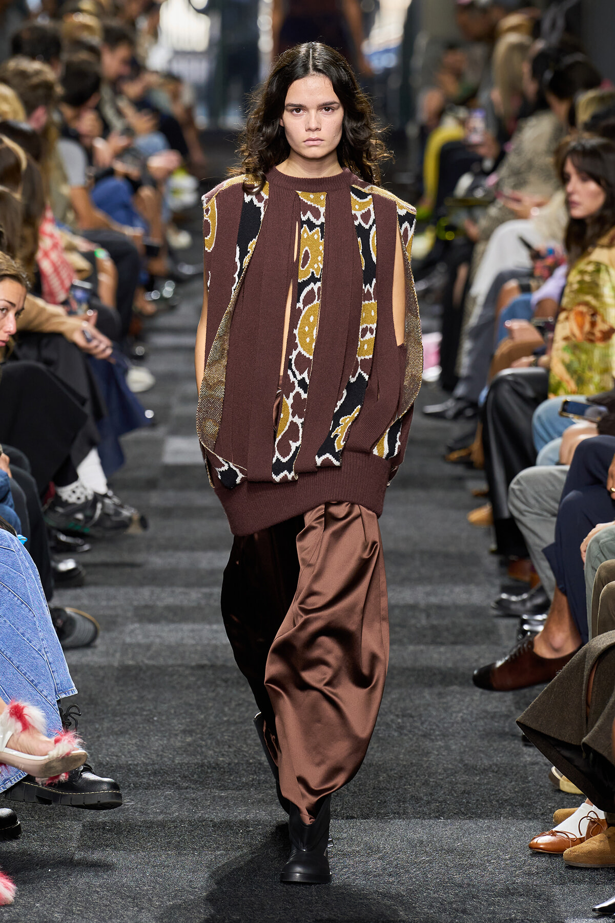 Model walks a fashion runway in a brown patterned knit sweater with cutout sleeves and loose satin brown pants, audience on both sides watching.