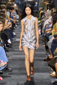 Model walking a runway in a white crochet lattice mini dress with cutouts and brown ankle boots.
