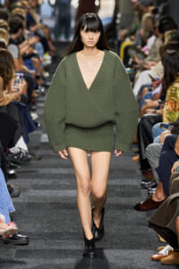 Model walks a runway wearing an oversized olive-green knit sweater with a deep V-neck and a matching mini skirt, black ankle boots on.