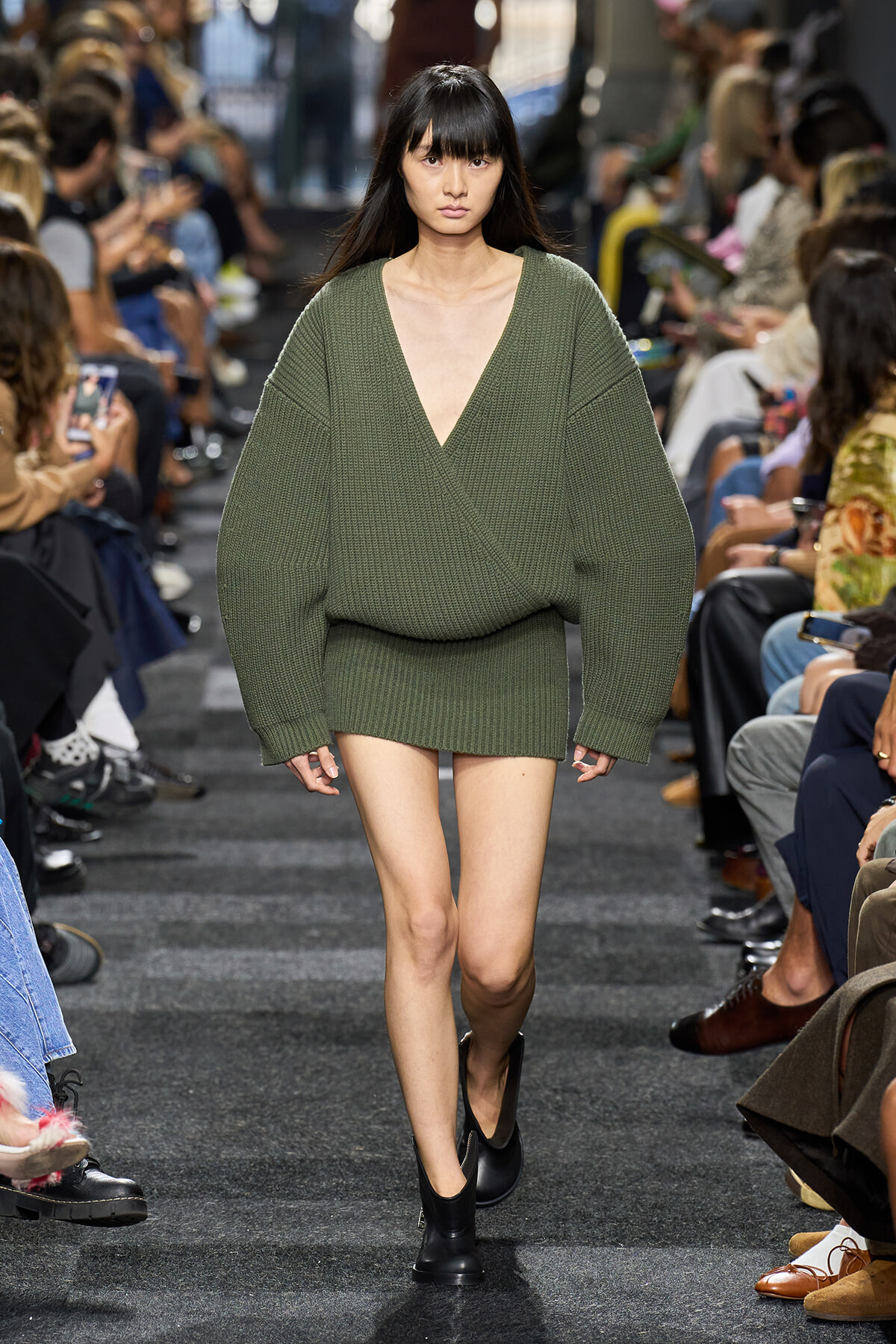 Model walks a runway wearing an oversized olive-green knit sweater with a deep V-neck and a matching mini skirt, black ankle boots on.