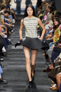 Model walks the runway in a cream sleeveless top with printed text and a sculptural black flared skirt, black ankle boots, audience on both sides of the catwalk.