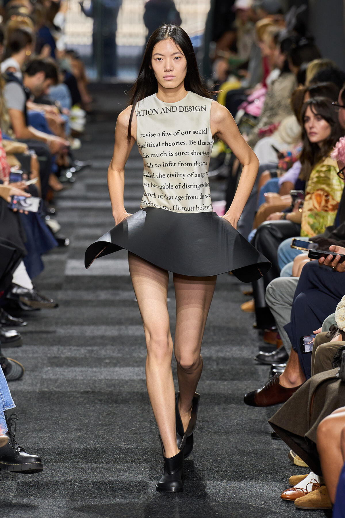 Model walks the runway in a cream sleeveless top with printed text and a sculptural black flared skirt, black ankle boots, audience on both sides of the catwalk.