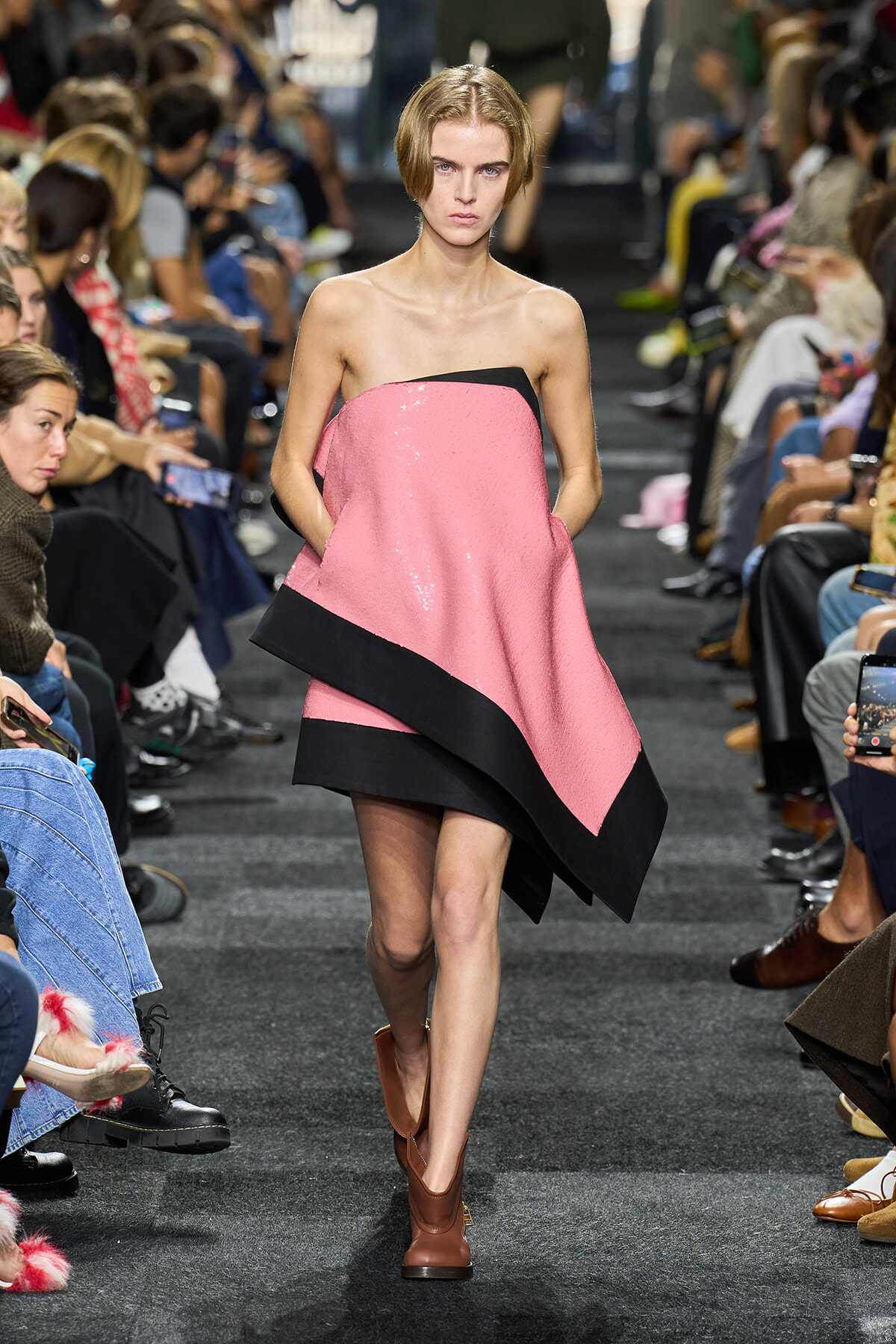 Model walks the runway in a pink strapless dress with a black hem, paired with brown ankle boots.