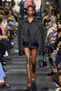 Model walks the runway in an oversized black jacket, black mini skirt, and brown ankle boots, carrying a small black handbag.