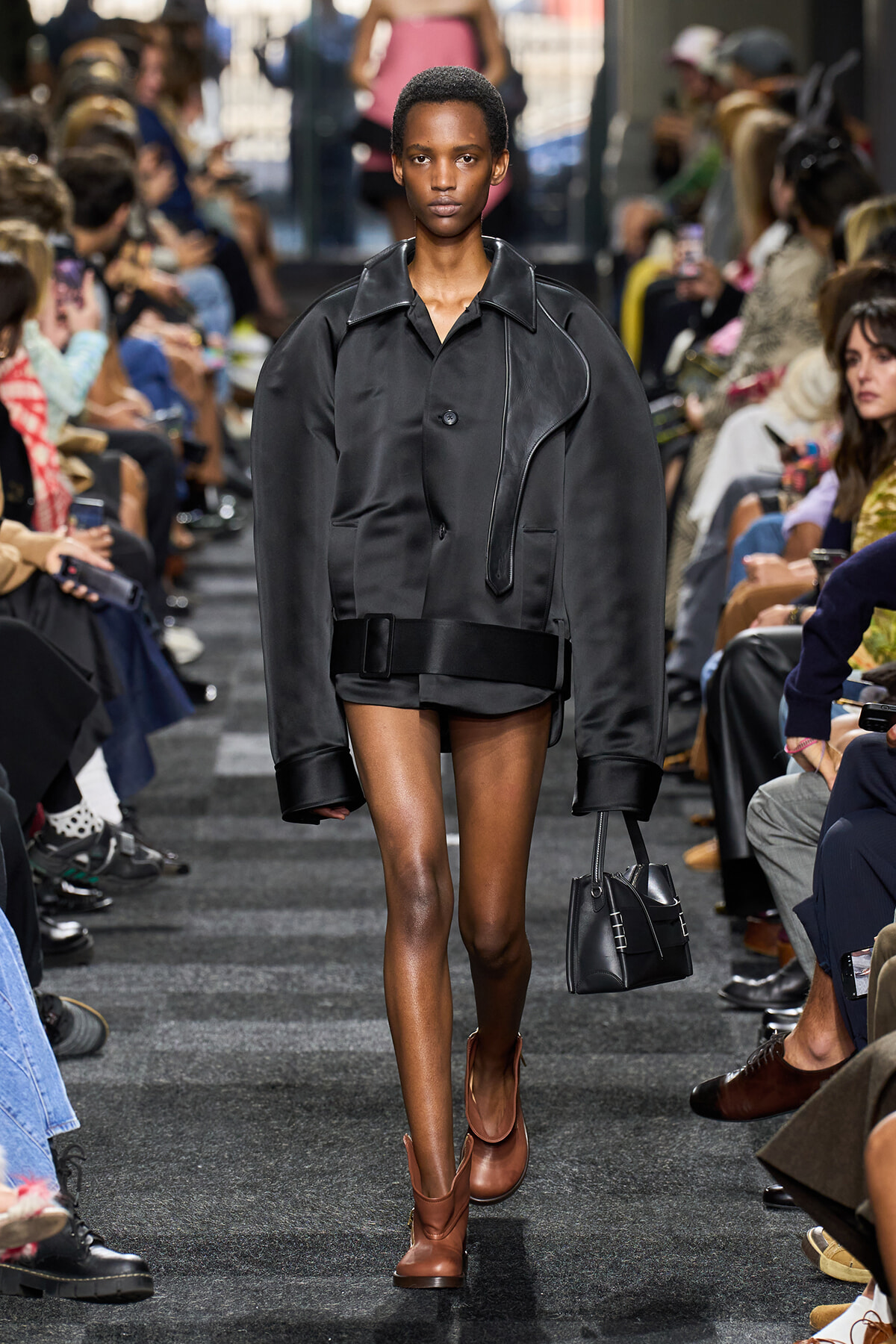 Model walks the runway in an oversized black jacket, black mini skirt, and brown ankle boots, carrying a small black handbag.