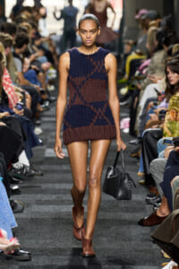 Model walking a fashion show runway in a navy and brown knit sleeveless mini dress, carrying a black handbag.