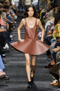 Model walking a runway in a brown sleeveless leather dress with a large circular, flared skirt, paired with black ankle boots.