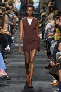 Model walks the runway in a sleeveless brown knit V-neck mini dress with a white top underneath, paired with brown ankle boots, audience on both sides.