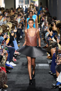 Runway model wearing a brown sleeveless leather top and a large black flared skirt, as photographers line the catwalk.