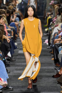 Female model walks a runway in a mustard sleeveless dress with cut slits, a cream and orange scarf tied around her leg, and brown ankle boots amid a seated audience.