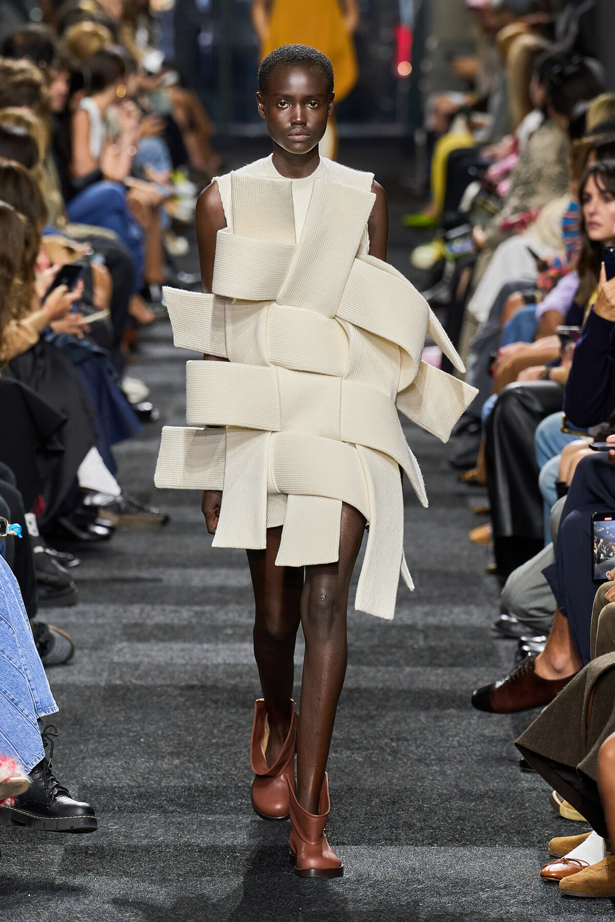 Black female model walks a runway in a sculptural off-white dress made of wide fabric bands wrapped around the torso and hips, paired with brown ankle boots.